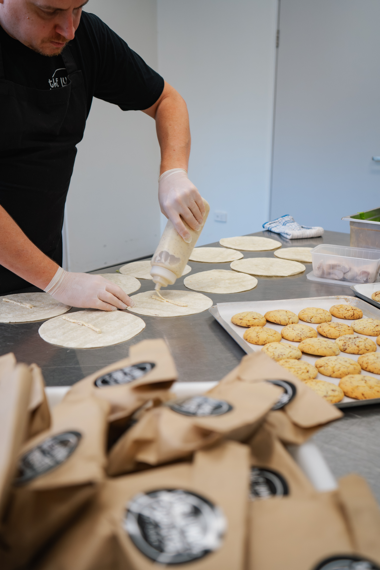 Chef assembling fresh wraps alongside trays of cookies and packed Lunch Project bags